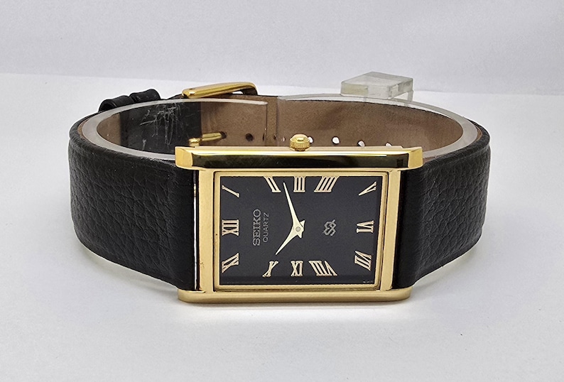 May include: A rectangular gold-toned Seiko Quartz watch with a black face and Roman numeral hour markers. The watch has a black leather band and a gold-toned buckle. The watch face displays the Seiko logo and the word "QUARTZ".