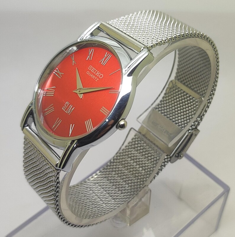 Puede incluir: Un reloj Seiko Quartz plateado con una esfera roja brillante y n&uacute;meros romanos. El reloj tiene una correa de malla. La palabra "SLIM" est&aacute; impresa en la esfera. El reloj se exhibe en un soporte transparente.