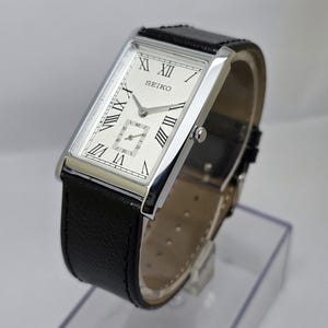 May include: A rectangular Seiko watch with a silver face, black Roman numerals, and a small seconds dial. The watch has a stainless steel case and a black leather strap. The watch is displayed at an angle.