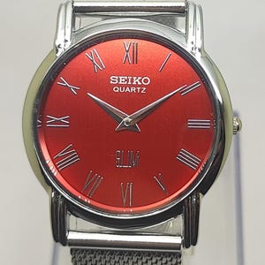 Puede incluir: Reloj Seiko Quartz plateado con esfera roja y agujas plateadas. El reloj presenta n&uacute;meros romanos y una correa de malla. La palabra "SLIM" est&aacute; impresa en la esfera.