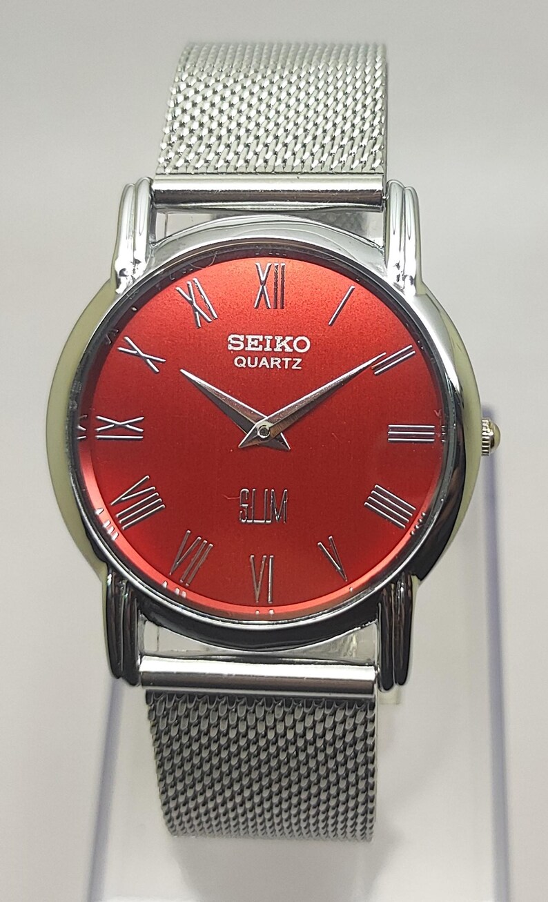 Puede incluir: Reloj Seiko Quartz plateado con esfera roja y agujas plateadas. El reloj presenta n&uacute;meros romanos y una correa de malla. La palabra "SLIM" est&aacute; impresa en la esfera.