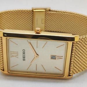 May include: A gold Seiko wristwatch with a rectangular face and a mesh strap. The watch face is cream-coloured with gold hands and markers, and the date is displayed at the 3 o'clock position. The strap is a woven mesh design.