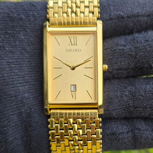 Puede incluir: Un reloj Seiko dorado con una esfera rectangular y una correa dorada a juego. La esfera del reloj muestra el logotipo de Seiko, números romanos y una ventana de fecha. El reloj es de diseño clásico, adecuado para ocasiones formales.