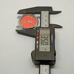 Puede incluir: Un reloj plateado con esfera naranja y n&uacute;meros romanos se mide con un calibrador digital. El calibrador muestra una medida de 8,89 cm. El reloj es un Seiko Quartz.