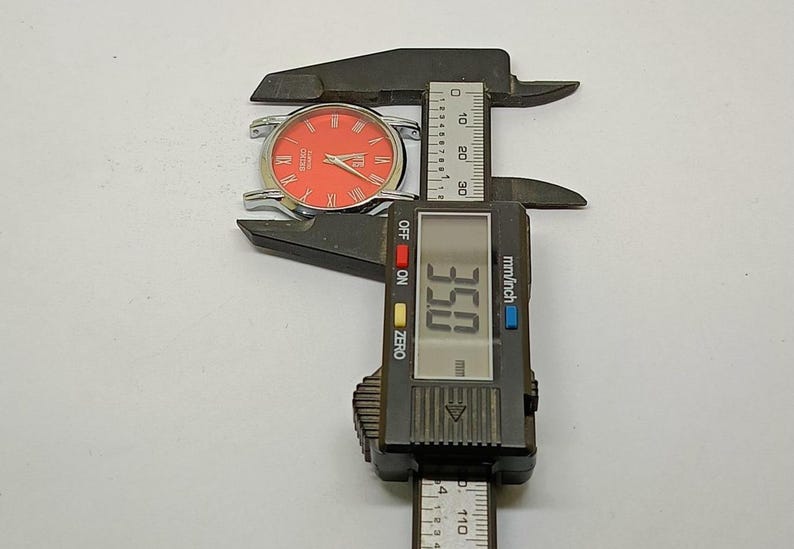 Puede incluir: Un reloj plateado con esfera naranja y n&uacute;meros romanos se mide con un calibrador digital. El calibrador muestra una medida de 8,89 cm. El reloj es un Seiko Quartz.