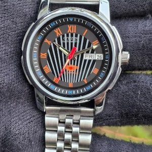 May include: Silver-toned Citizen automatic watch with a black and orange dial. The watch features Roman numerals, a day/date display, and a silver link bracelet. The dial has a striped design with orange accents and red hands. The watch is displayed on a dark surface.