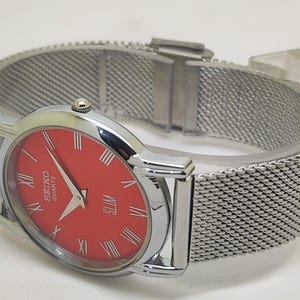 Puede incluir: Un reloj Seiko Quartz plateado con esfera roja y n&uacute;meros romanos. El reloj presenta una correa de metal de malla y un perfil delgado. Un reloj cl&aacute;sico.