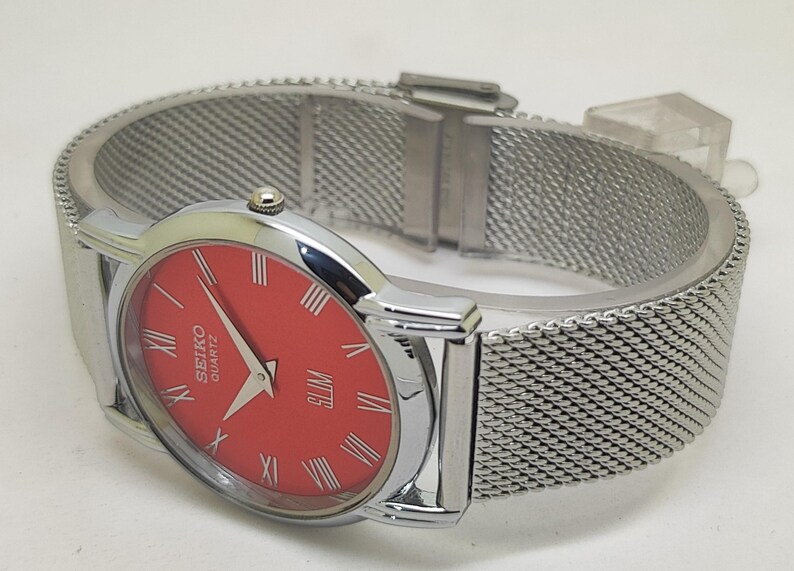 Puede incluir: Un reloj Seiko Quartz plateado con esfera roja y n&uacute;meros romanos. El reloj presenta una correa de metal de malla y un perfil delgado. Un reloj cl&aacute;sico.