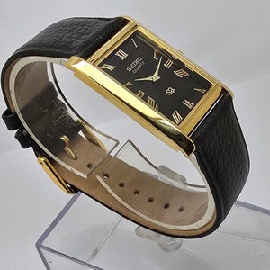 May include: A rectangular Seiko Quartz watch with a black face and gold-tone case. The watch features Roman numeral hour markers and a black leather band. The watch is displayed on a clear plastic stand.