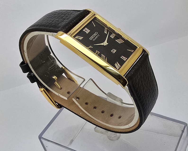 May include: A rectangular Seiko Quartz watch with a black face and gold-tone case. The watch features Roman numeral hour markers and a black leather band. The watch is displayed on a clear plastic stand.