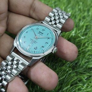 May include: A vintage HMT Janata wristwatch with a light blue dial and Arabic numerals. The watch has a silver-coloured stainless steel band and case. The dial features the text "HMT Janata" and "Para Shock 17 Jewels".