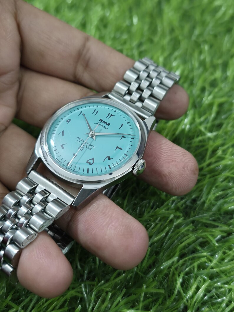 May include: A vintage HMT Janata wristwatch with a light blue dial and Arabic numerals. The watch has a silver-coloured stainless steel band and case. The dial features the text "HMT Janata" and "Para Shock 17 Jewels".