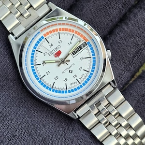 May include: A silver Seiko automatic wristwatch with a white face, featuring blue and orange accents. The watch displays the day and date, with a stainless steel band. The watch face includes the text "SEIKO" and "AUTOMATIC".