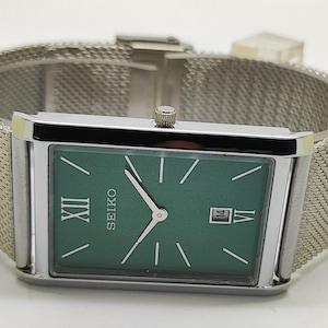 Puede incluir: Reloj Seiko rectangular con esfera verde y agujas plateadas. El reloj tiene una correa de malla plateada y una pantalla de fecha a las 3 en punto. La esfera del reloj presenta el logotipo de Seiko y el número romano XII. Diseño clásico.