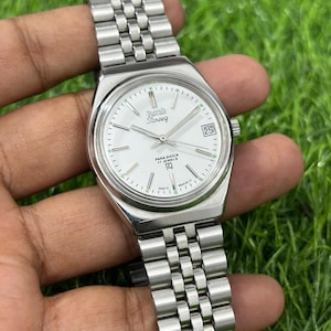 Op de afbeelding: Zilverkleurig polshorloge met een witte wijzerplaat, met de merknaam "Tareeq" en een datumweergave. Het horloge heeft een metalen band en bevindt zich tegen een groene achtergrond. De wijzerplaat bevat de tekst "Para Shock 17 Jewels".