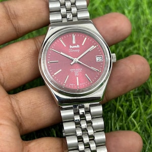 May include: A silver HMT Tareeq wristwatch with a red face. The watch has a date window and a stainless steel strap. The watch face displays the text "hmt Tareeq" and "PARA SHOCK 17 JEWELS". The watch is a classic design.