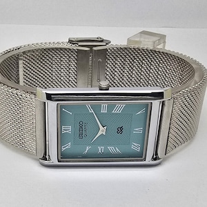 Puede incluir: Un reloj Seiko Quartz rectangular con una correa de malla plateada. La esfera del reloj es de color verde azulado claro con números romanos y agujas plateadas. La caja y la correa son de acero inoxidable.