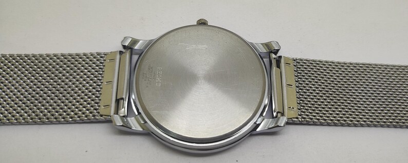 Puede incluir: Un reloj de pulsera plateado con una correa de malla. La caja del reloj es redonda con una parte trasera plateada y un bisel plateado. La correa de malla es de color plata clara y est&aacute; unida a la caja del reloj con conectores plateados y dorados.