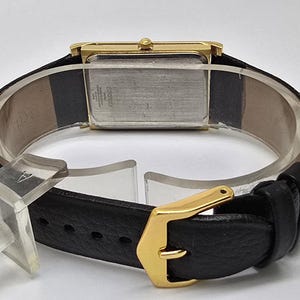 May include: A gold-toned rectangular wristwatch with a black leather band. The watch has a gold buckle and a light-colored watch face. The back of the watch is visible, showing the metal casing and some text.