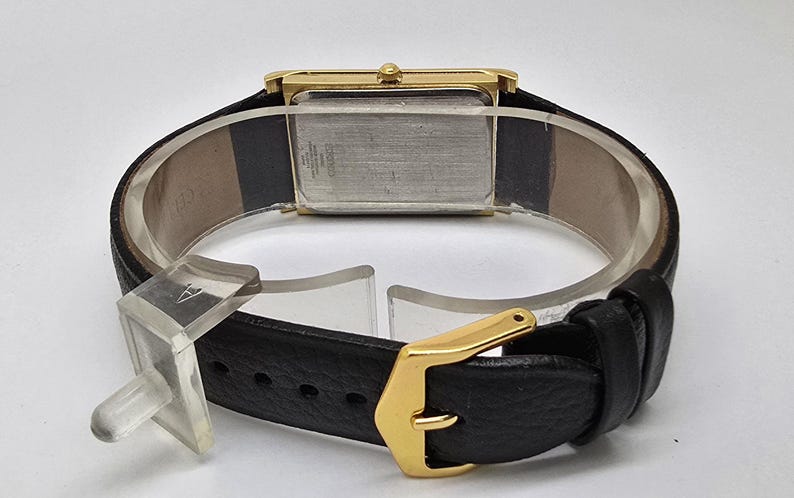 May include: A gold-toned rectangular wristwatch with a black leather band. The watch has a gold buckle and a light-colored watch face. The back of the watch is visible, showing the metal casing and some text.