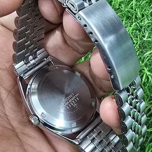 Puede incluir: Un reloj Seiko de acero inoxidable con una correa plateada. La esfera del reloj no es visible, pero la parte trasera del reloj es visible, con el texto "SEIKO WATER RESISTANT 335718 TL". El reloj se sostiene sobre un fondo de hierba verde.