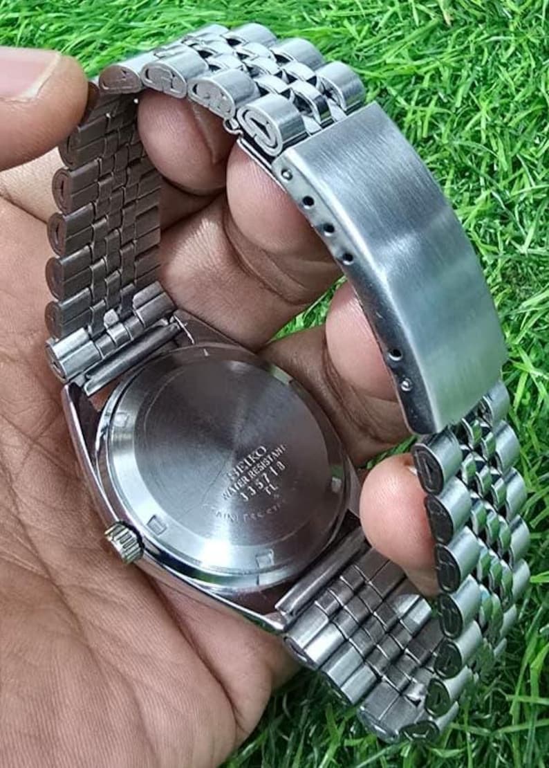 Puede incluir: Un reloj Seiko de acero inoxidable con una correa plateada. La esfera del reloj no es visible, pero la parte trasera del reloj es visible, con el texto "SEIKO WATER RESISTANT 335718 TL". El reloj se sostiene sobre un fondo de hierba verde.