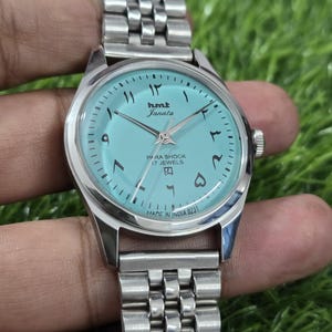 May include: A silver wristwatch with a light blue face and Arabic numerals. The watch face displays the brand name "hmt Janata" and the text "PARA SHOCK 17 JEWELS". The watch band is made of linked metal. The text "MADE IN INDIA 0231" is visible.