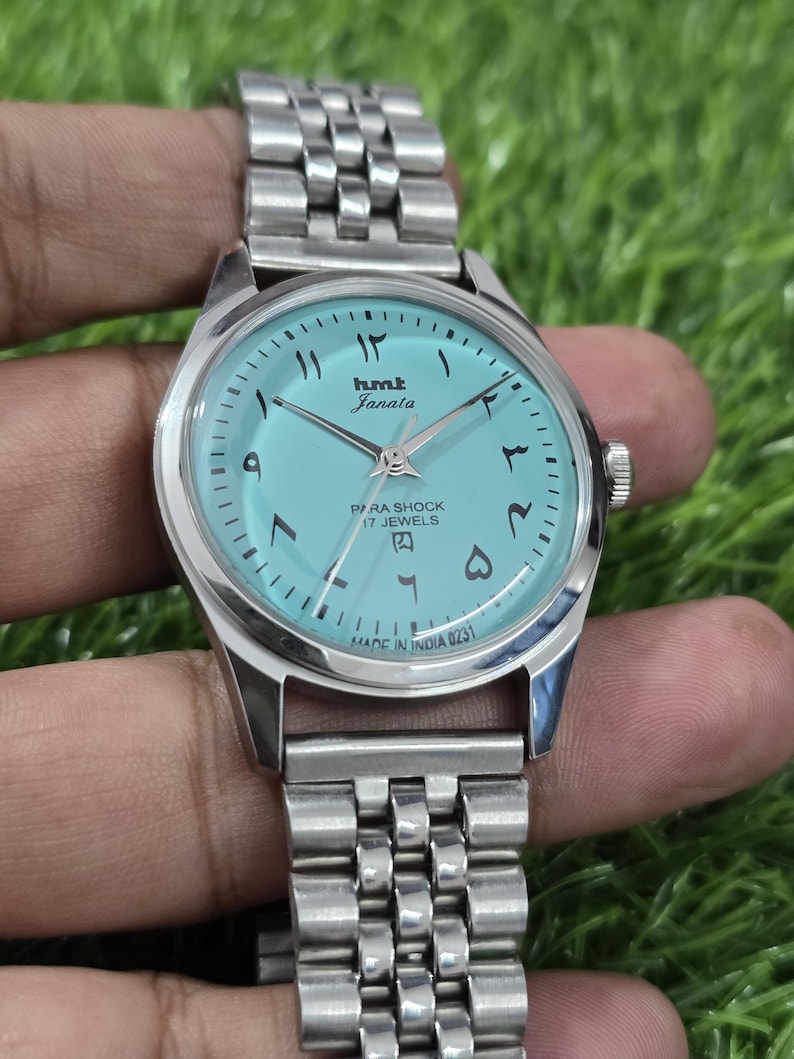 May include: A silver wristwatch with a light blue face and Arabic numerals. The watch face displays the brand name "hmt Janata" and the text "PARA SHOCK 17 JEWELS". The watch band is made of linked metal. The text "MADE IN INDIA 0231" is visible.