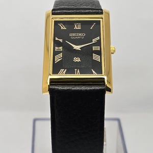 May include: A rectangular gold-toned Seiko Quartz watch with a black face and black leather band. The watch face features gold Roman numerals and hands. The watch has a small gold crown and a black leather band.