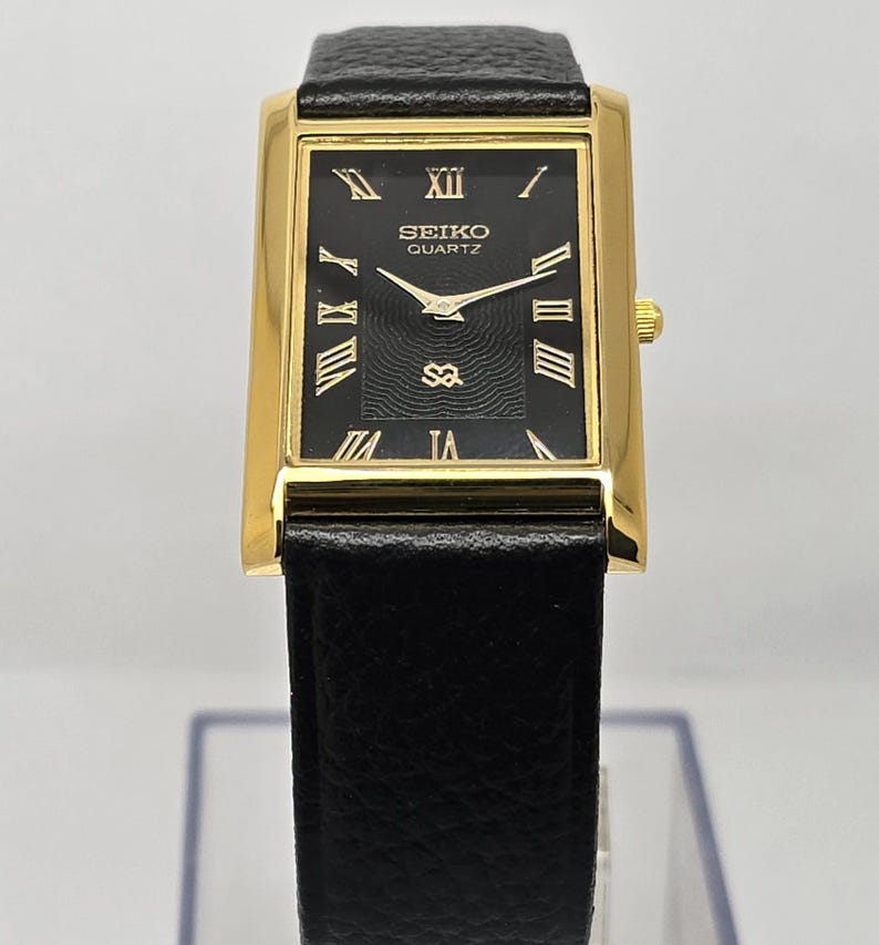 May include: A rectangular gold-toned Seiko Quartz watch with a black face and black leather band. The watch face features gold Roman numerals and hands. The watch has a small gold crown and a black leather band.