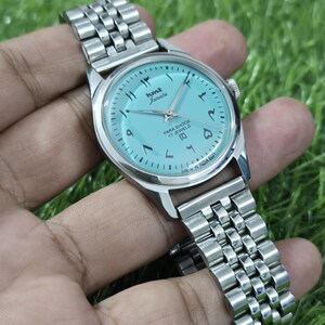 May include: A silver wristwatch with a light blue face and a metal strap. The watch face displays Arabic numerals and the text "hmt Janata PARA SHOCK 17 JEWELS". The watch is held against a green background.