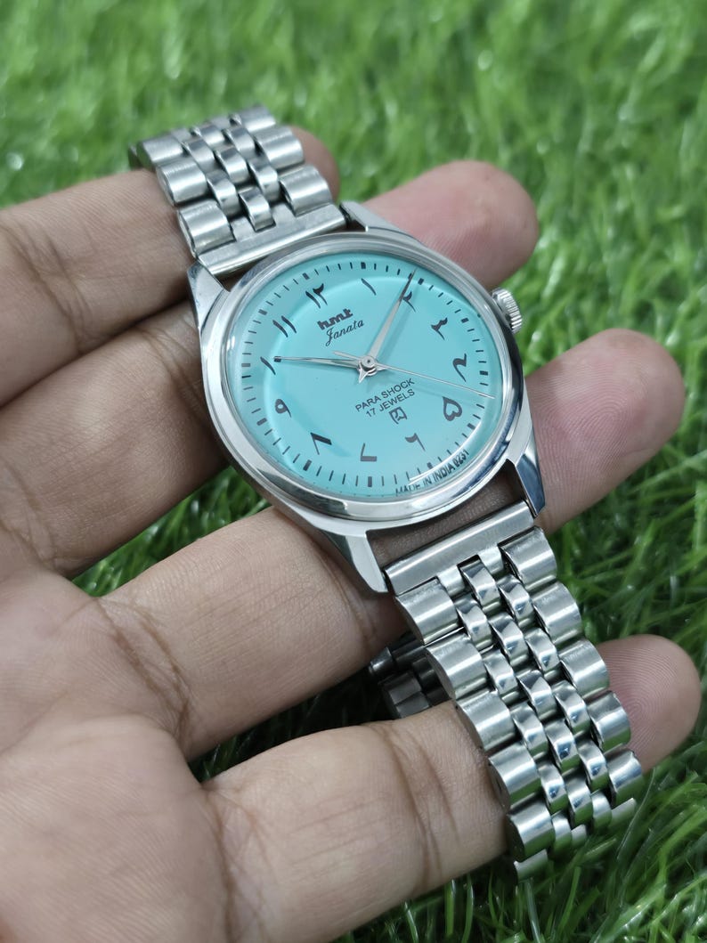May include: A silver wristwatch with a light blue face and a metal strap. The watch face displays Arabic numerals and the text "hmt Janata PARA SHOCK 17 JEWELS". The watch is held against a green background.