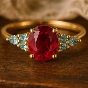 May include: A gold ring featuring a large oval-cut ruby center stone, flanked by smaller, light blue pear-shaped gemstones. The ring is set on a wooden surface, showcasing the vibrant red and blue hues.