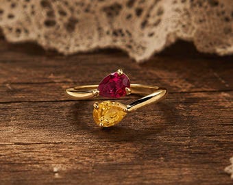 May include: A gold ring featuring two pear-shaped gemstones, one red and one yellow. The ring's band curves around the gemstones, set against a rustic wooden surface. The gemstones are the focal point, with a delicate lace detail in the background.