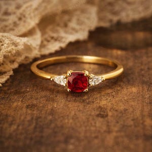 Op de afbeelding: Een gouden ring met een vierkante, dieprode middensteen en twee kleinere, driehoekige witte stenen aan weerszijden. De ring ligt op een donkerhouten oppervlak met een zachte, crèmekleurige stof op de achtergrond.