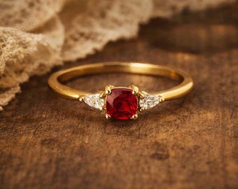 May include: A gold ring featuring a square, deep red gemstone at its centre, flanked by two smaller, triangular white stones. The ring is displayed on a dark wooden surface, with a cream-coloured fabric in the background.