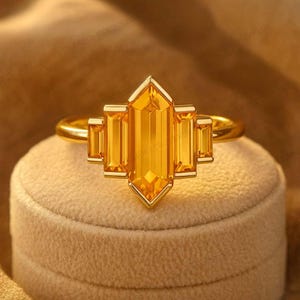 Könnte beinhalten: Ein goldener Ring mit einem großen, rechteckigen, gelben Edelstein und kleineren rechteckigen Steinen. Der Ring wird auf einer beigen Samtoberfläche präsentiert, die sein Design und seine Farbe hervorhebt.