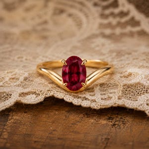 May include: A gold ring featuring a central oval-cut deep red gemstone held by four prongs. The band has a smooth, polished finish and a V-shaped design that tapers towards the centre setting, showcasing a classic and elegant jewellery piece.