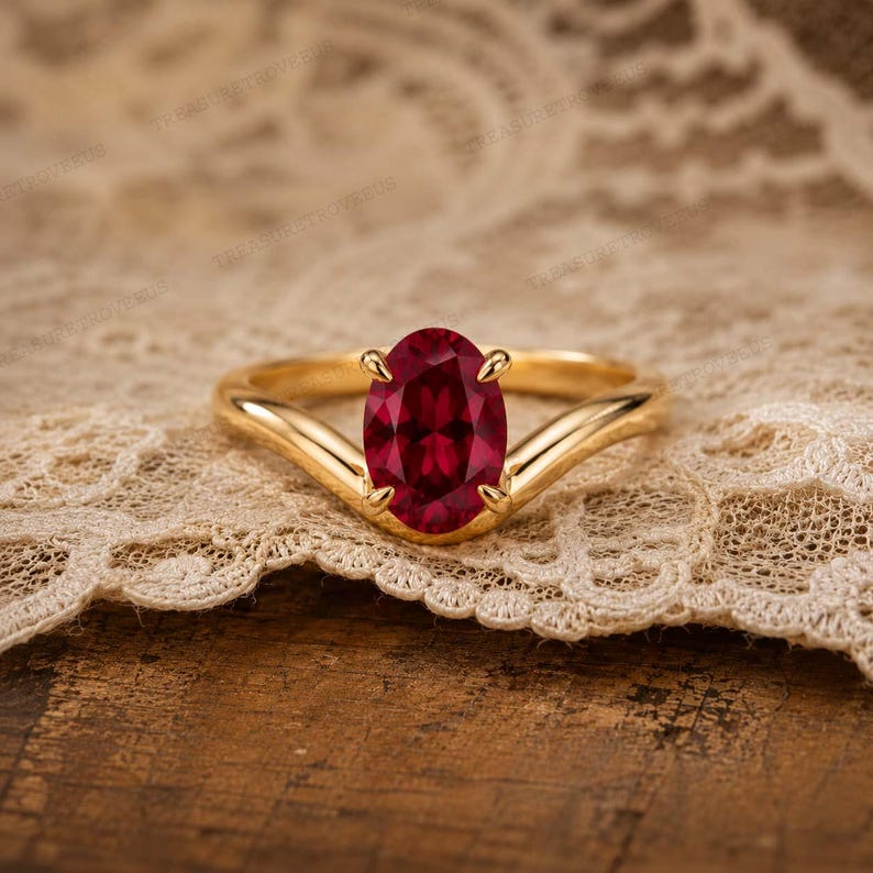 May include: A gold ring featuring a central oval-cut deep red gemstone held by four prongs. The band has a smooth, polished finish and a V-shaped design that tapers towards the centre setting, showcasing a classic and elegant jewellery piece.
