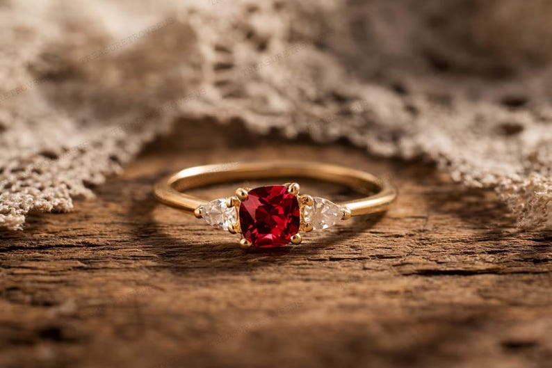 Puede incluir: Un anillo de oro con una piedra preciosa roja oscura de talla coj&iacute;n como piedra central, flanqueada por dos piedras preciosas claras m&aacute;s peque&ntilde;as en forma de pera. El anillo descansa sobre una superficie de madera, con tela de encaje en el fondo.