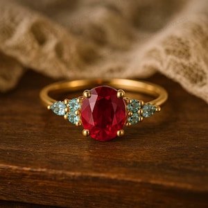 K&ouml;nnte beinhalten: Ein goldener Ring mit einem gro&szlig;en, oval geschliffenen Rubin als Mittelstein, flankiert von kleineren, hellblauen, birnenf&ouml;rmigen Edelsteinen. Der Ring liegt auf einer Holzoberfl&auml;che, die die leuchtenden Rot- und Blaut&ouml;ne zeigt.