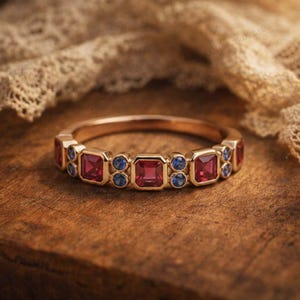 May include: A gold ring featuring a series of alternating square-cut red gemstones and pairs of small round blue gemstones set along the band. The metal band has a polished finish, and the stones are arranged in a repeating pattern around the front.