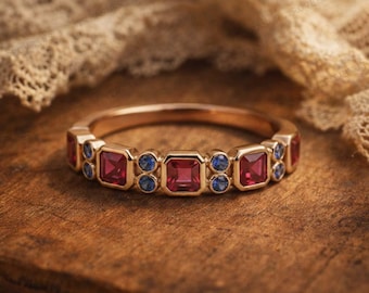 May include: A gold ring featuring a series of alternating square-cut red gemstones and pairs of small round blue gemstones set along the band. The metal band has a polished finish, and the stones are arranged in a repeating pattern around the front.