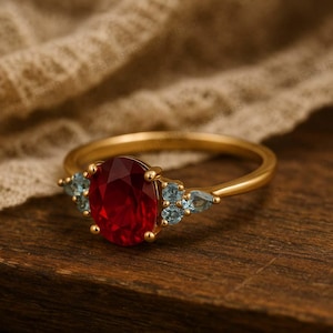 K&ouml;nnte beinhalten: Ein goldener Ring mit einem gro&szlig;en, oval geschliffenen roten Edelstein als Mittelpunkt. Auf beiden Seiten befinden sich kleinere, hellblaue Edelsteine. Der Ring liegt auf einer Holzoberfl&auml;che, die das Design und den Farbkontrast hervorhebt.