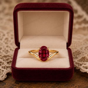 May include: A gold ring featuring a central oval-cut deep red gemstone held by four prongs, set within a dark red velvet jewellery box with a cream-coloured interior lining. The ring band has a smooth, polished finish and a V-shaped design.