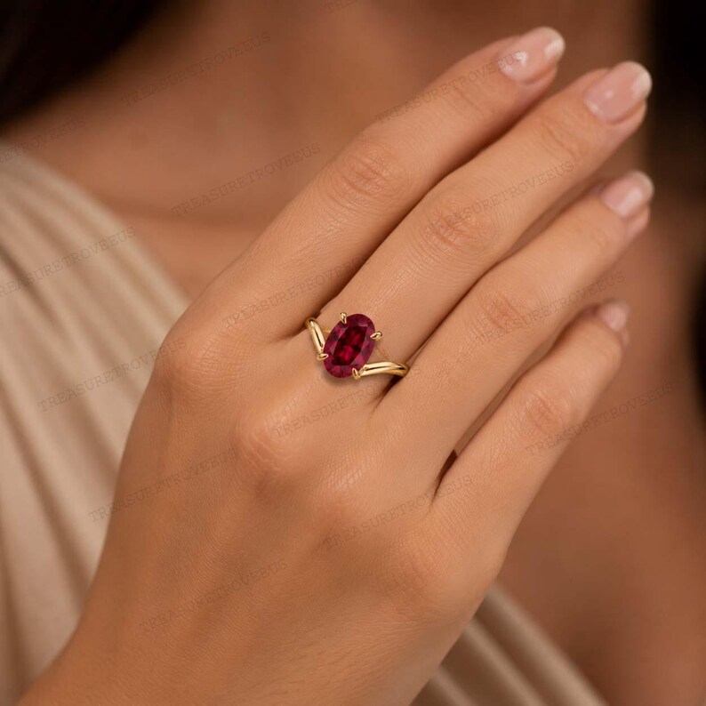 May include: A gold ring featuring a central oval-cut deep red gemstone held by four prongs. The band has a V-shaped design that tapers towards the stone, creating a pointed setting. The metal has a polished yellow finish, highlighting the contrast.
