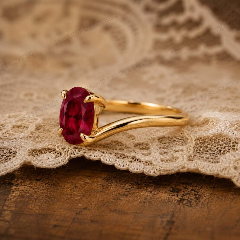 May include: A gold ring featuring a prominent oval-cut deep red gemstone held by four prongs. The band has a smooth, polished finish and a slightly curved, bypass-style design that tapers as it extends from the central stone setting.