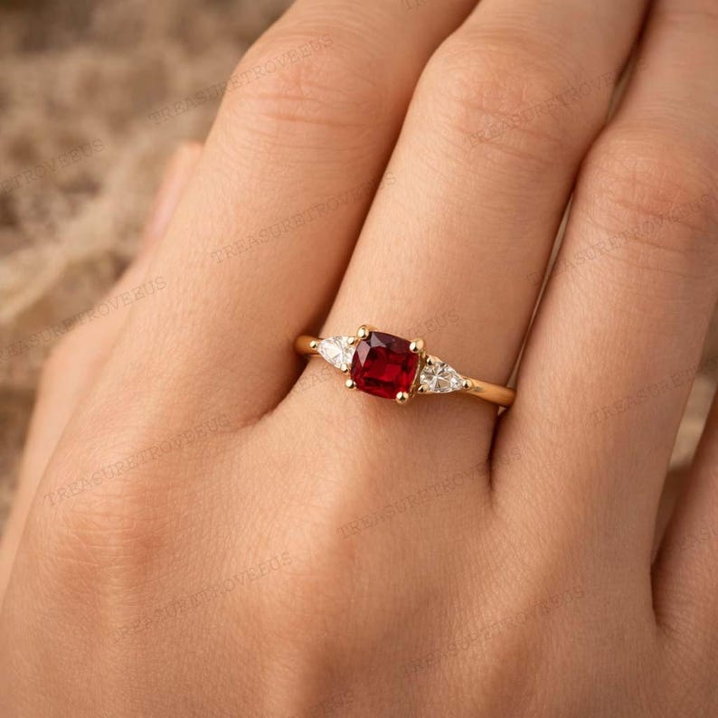 Puede incluir: Un anillo de oro con una piedra preciosa roja cuadrada en el centro, flanqueada por dos piedras preciosas blancas triangulares m&aacute;s peque&ntilde;as. El anillo tiene una fina banda de oro pulido.