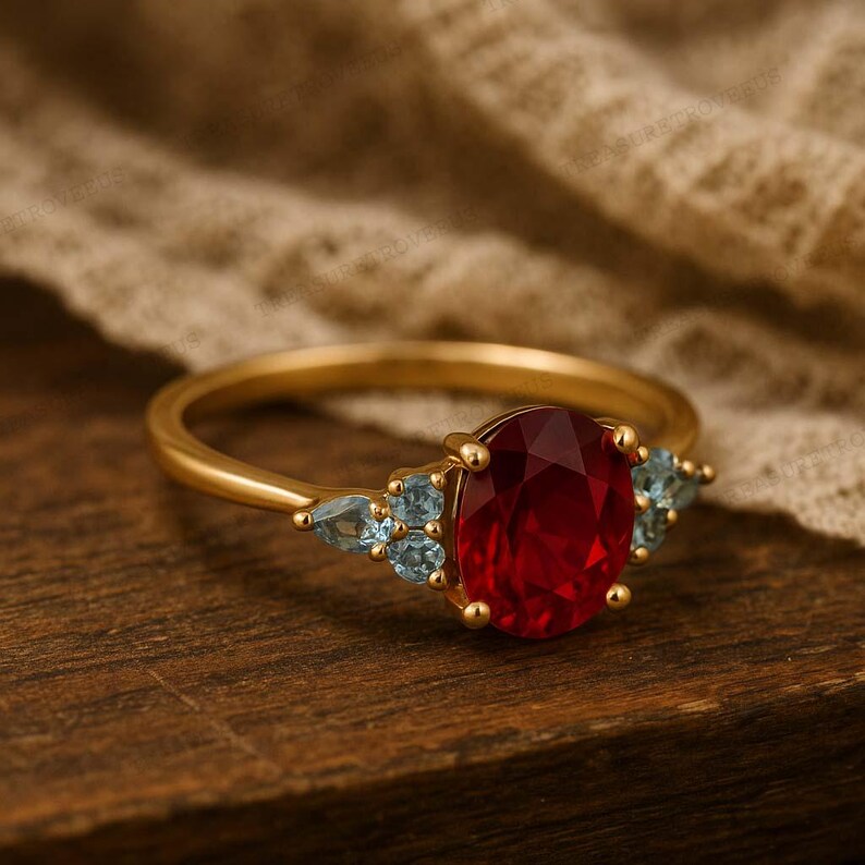 K&ouml;nnte beinhalten: Ein goldener Ring mit einem gro&szlig;en, oval geschliffenen, tiefroten Edelstein als Mittelpunkt. Flankiert wird er von kleineren, hellblauen Edelsteinen. Der Ring liegt auf einer Holzoberfl&auml;che und zeigt das Design des Schmuckst&uuml;cks.