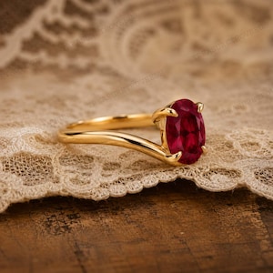 May include: A gold ring featuring a single oval-cut deep red gemstone held in a four-prong setting. The band has a smooth, polished finish and a curved design that tapers towards the centre stone, creating a sleek and refined appearance.
